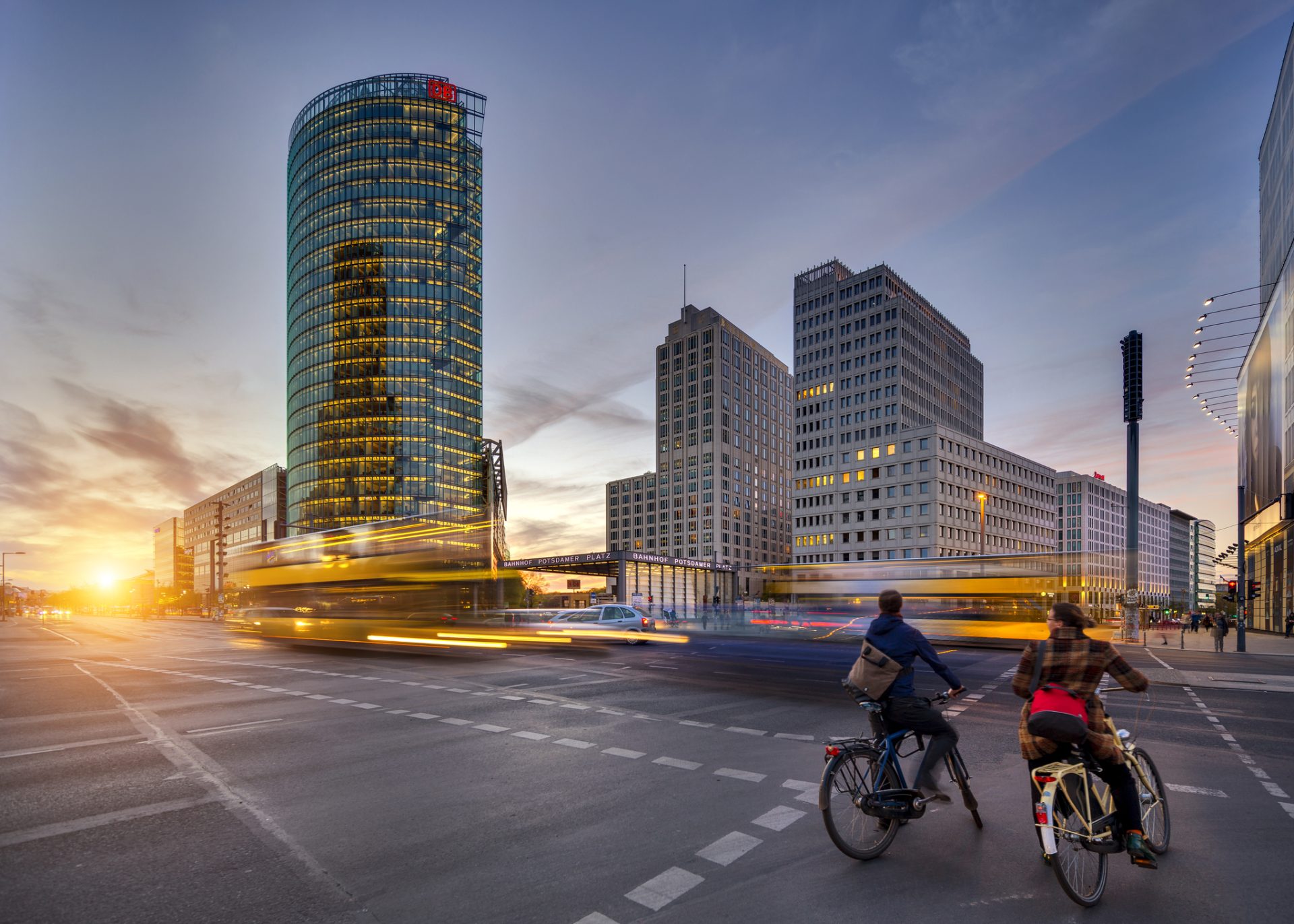 Two people on bicycles wait at a crosswalk in a city at sunset. Motion blur shows buses and cars moving through the intersection. Tall modern buildings are visible in the background.